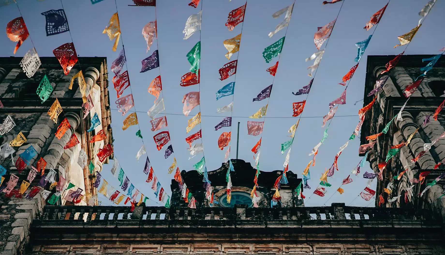 Colorful papel picado banners are strung across the sky above a historical stone building. The building features ornate architectural details, with a clear blue sky in the background, creating a festive and vibrant atmosphere.