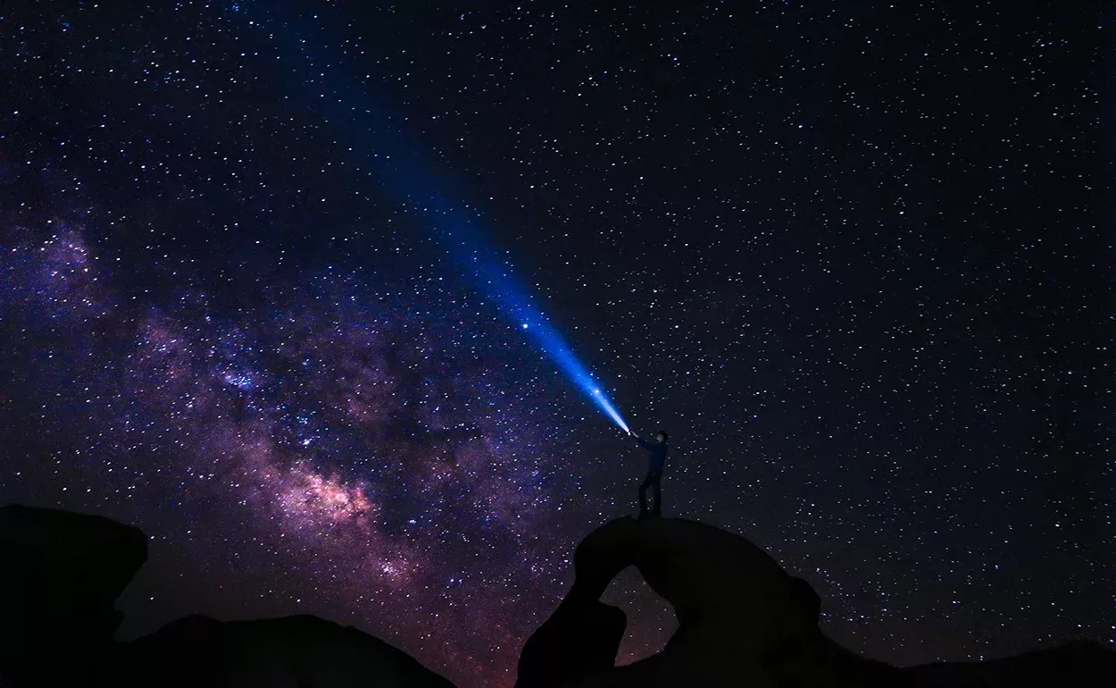Silhouette of a person standing on a rocky arch, pointing a flashlight toward a star-filled sky with the Milky Way visible. The beam creates a blue streak across the night sky, highlighting the celestial scene.