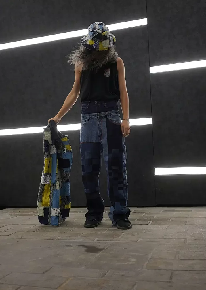 A person wearing a patchwork hat and jeans holds a matching patchwork jacket. They stand against a background with horizontal neon lights in a dark setting. The person has long hair and is wearing a sleeveless black shirt and sneakers.