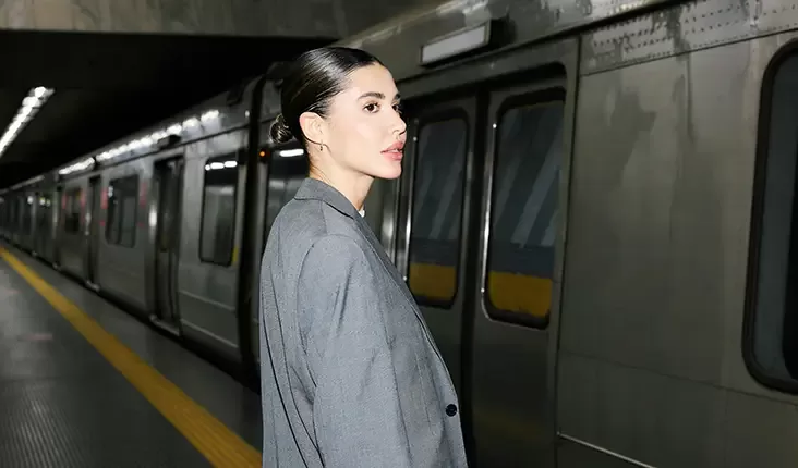 A woman with dark hair tied back stands on a subway platform, wearing a gray suit and staring thoughtfully into the distance. A subway train with closed doors is visible in the background.