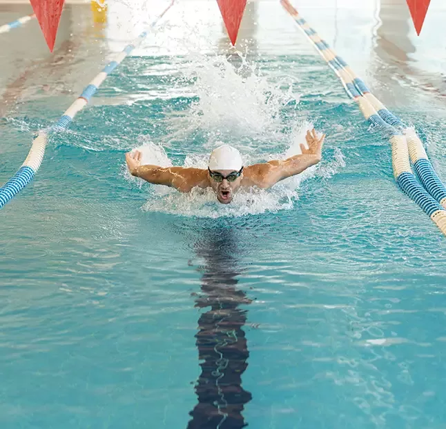 A swimmer in a white cap and goggles performs the butterfly stroke in a lane of an indoor swimming pool. Splashing water surrounds the swimmer as they extend their arms forward. Overhead, red triangular flags hang across the pool.