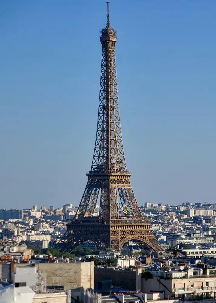 The Eiffel Tower in Paris, adorned with Olympic rings, stands tall against a clear blue sky. The cityscape below includes buildings and rooftops under the afternoon sun.
