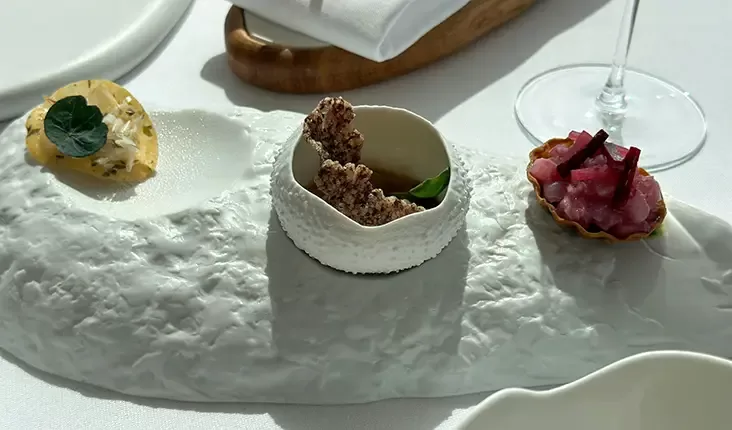 A white ceramic platter holds a small yellow mousse with a leaf garnish, a dish with a crunchy brown snack, and a tart filled with pink cubed pieces. A wine glass stem is visible in the background on a white tablecloth.