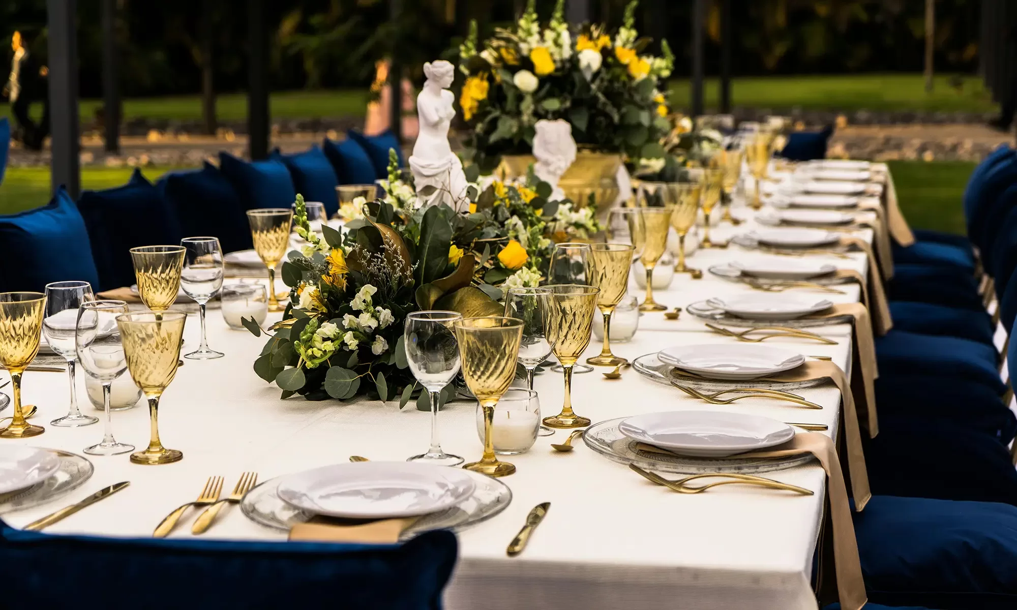 A long, elegantly set dining table outdoors, featuring gold-rimmed wine glasses, white plates with intricate patterns, and lush floral centerpieces with yellow and white flowers. Blue velvet chairs surround the table on a sunny day.