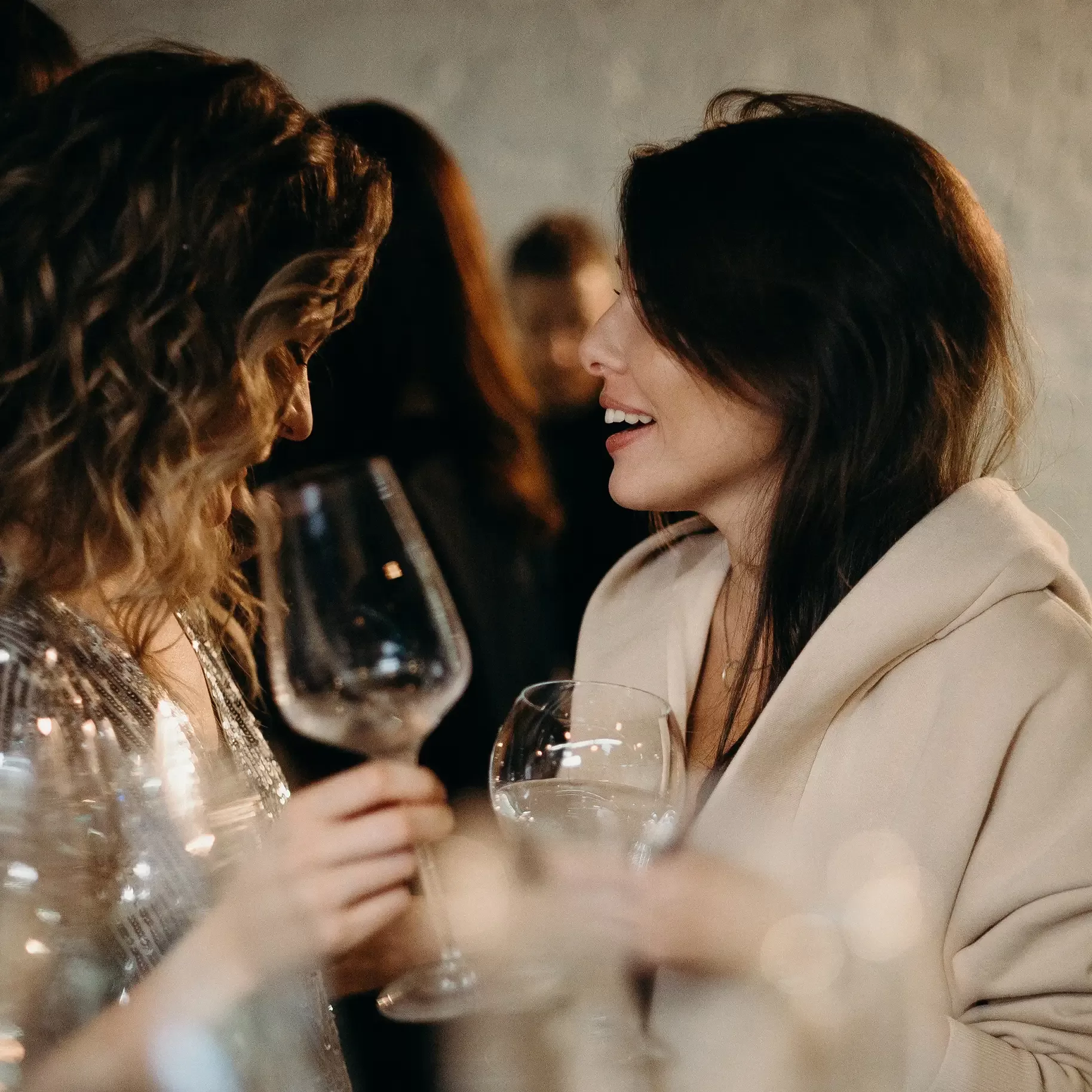 Two women at a social gathering hold empty wine glasses and smile at each other while talking. The background is softly blurred, highlighting their interaction.