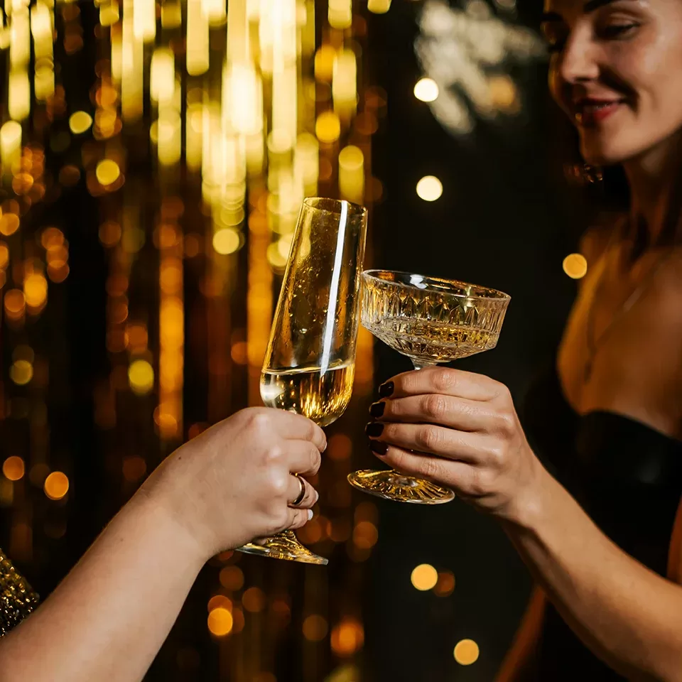 Two people toast with champagne flutes in front of golden, sparkling party decorations. One person wears black, and both hands hold elegant glasses filled with bubbly. The background glows with festive lights.