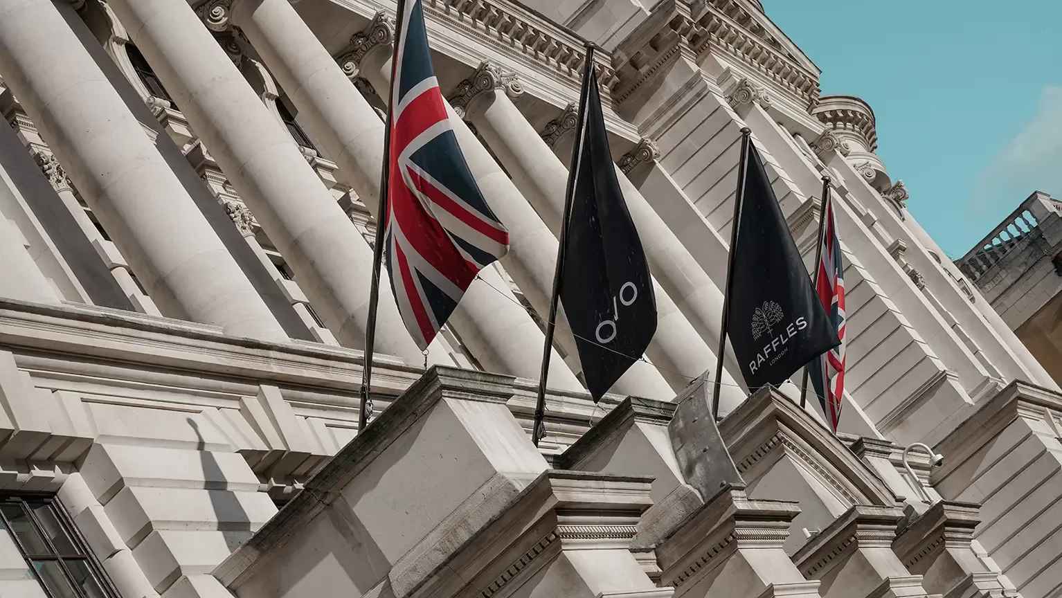 A close-up of a classical building façade with columns, featuring the Union Jack and three black flags, set against a clear blue sky.