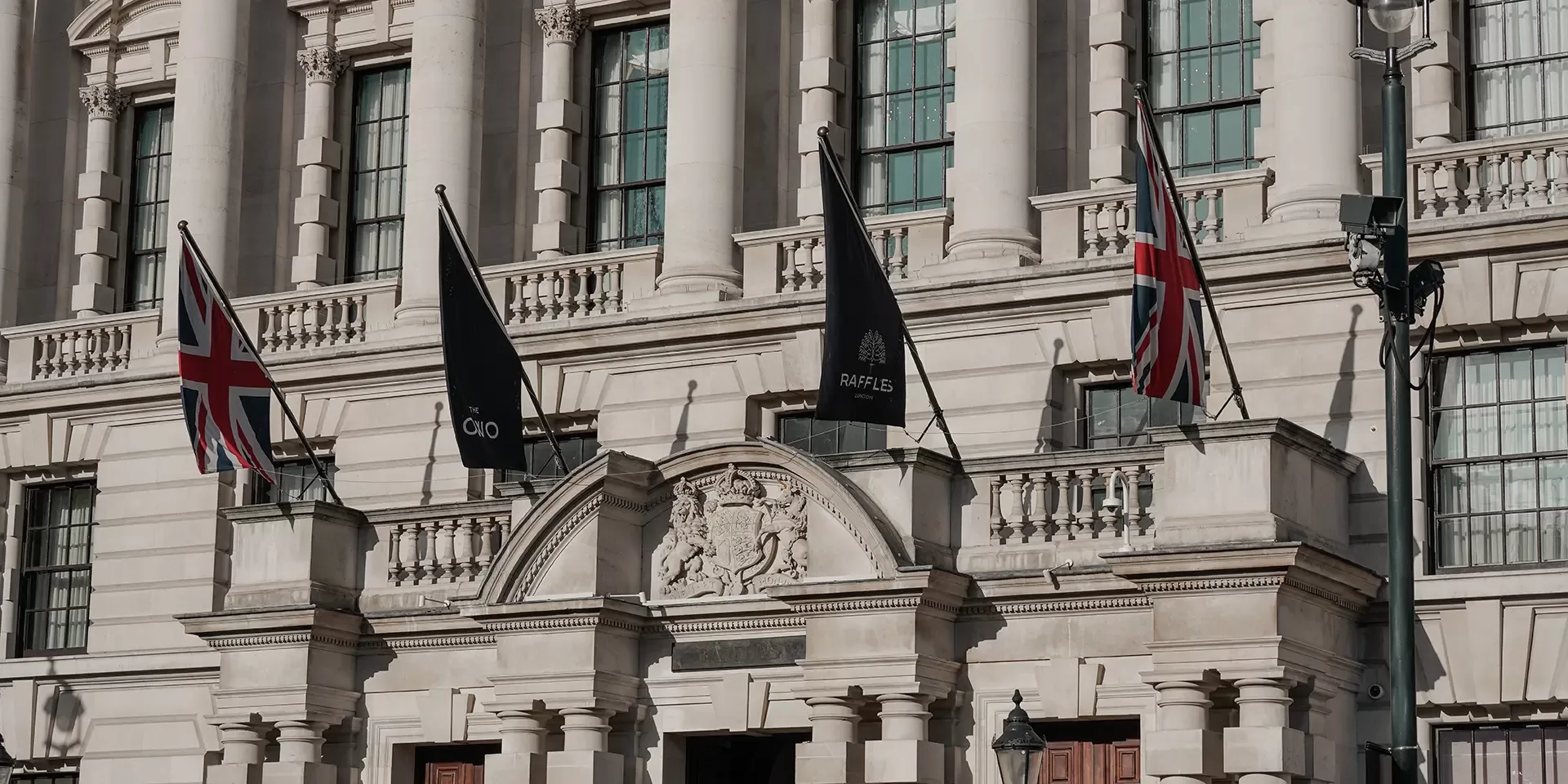 Historic stone building with columns and arched windows, featuring two British flags and two black flags that read Raffles above the entrance. Ornate crest and architectural details are visible above the doorways.