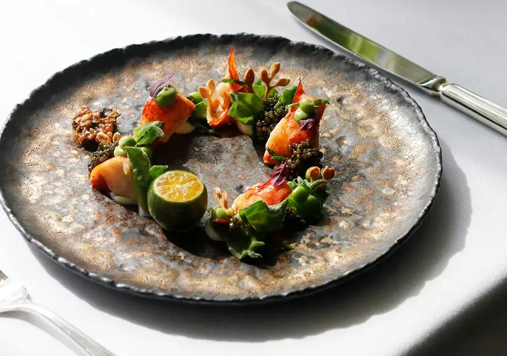A beautifully arranged gourmet dish on a textured, circular plate. It features vibrant ingredients including small seafood pieces, green leaves, delicate sauces, and a halved lime placed artistically in a circular pattern. A knife and fork are beside the plate.