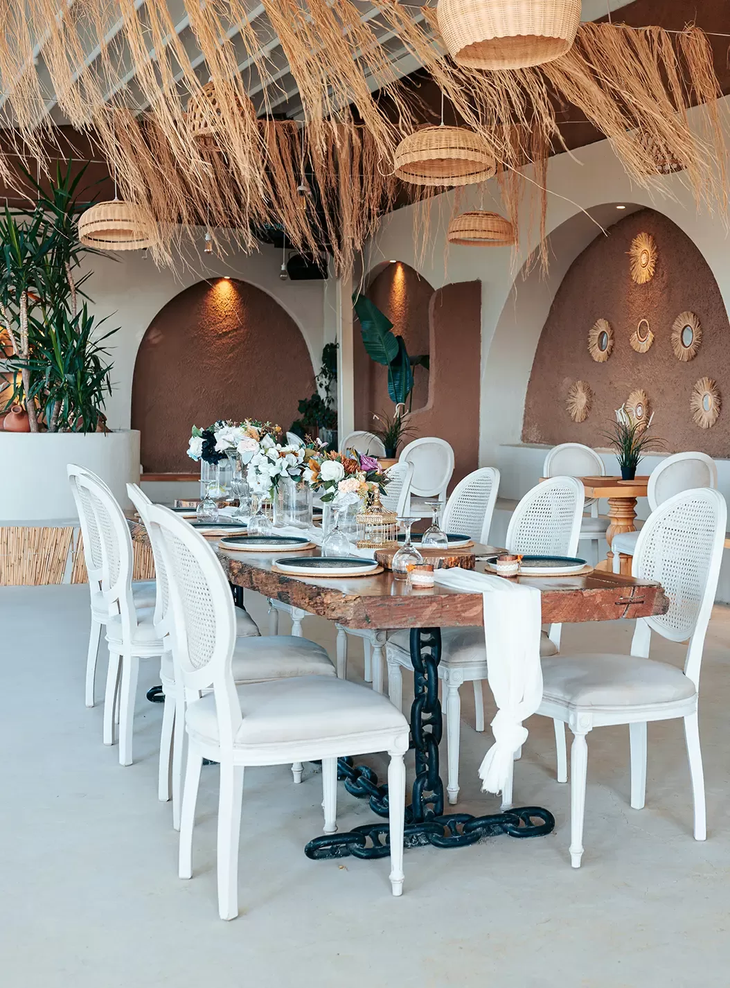 A rustic dining area features a wooden table with white chairs, surrounded by hanging wicker lights, dried grass decor, and arched walls with woven accents and plants. The table is set with plates, glasses, and floral arrangements.