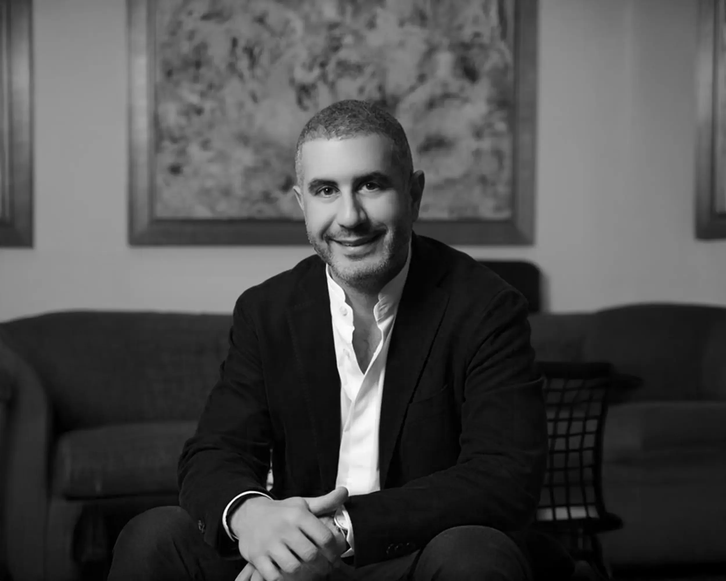 A man with short hair and a beard, dressed in a light shirt and dark blazer, sits smiling on a sofa in a living room. There is abstract artwork and a wire-frame chair in the background. The image is in black and white.