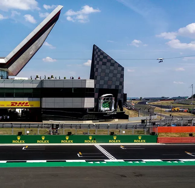 A view of the Silverstone Circuit finish line, with the modern pit building and observation tower in the background, and a helicopter flying in the sky above the race track. Rolex advertisements line the track barriers.