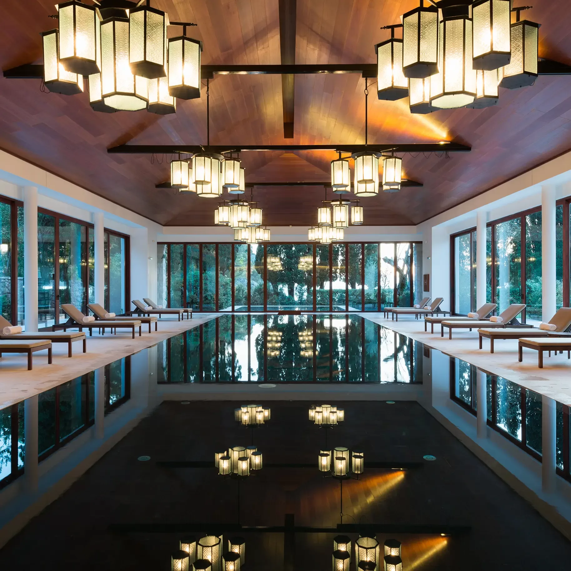 Luxurious indoor pool with lounge chairs along both sides, large windows letting in natural light, and modern hanging lanterns reflecting in the still water. Warm ambient lighting and wooden ceiling create a serene, inviting atmosphere.
