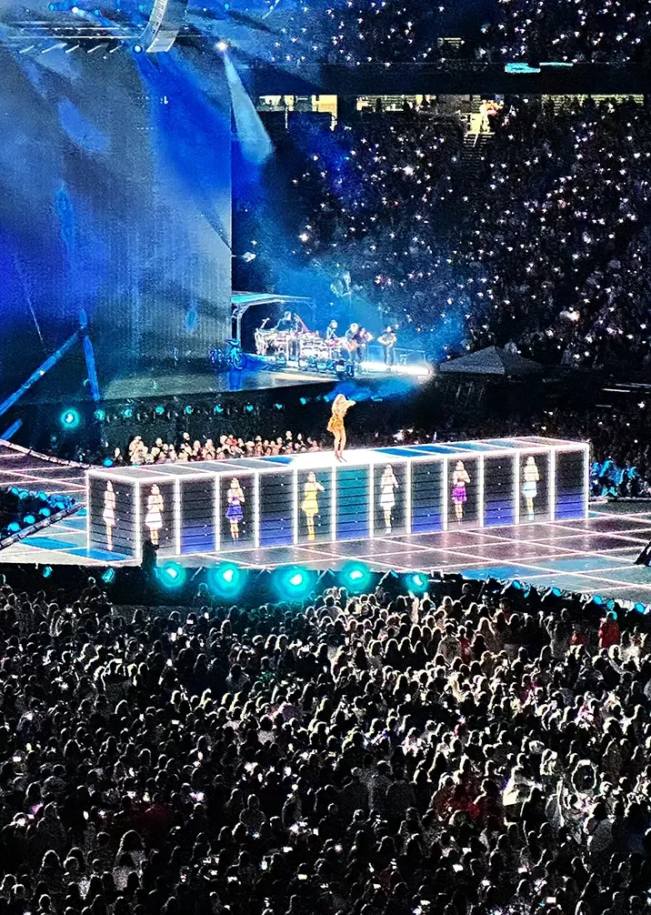 Taylor Swift on an illuminated stage in a packed stadium. The stage is elevated with bright lights and dancers positioned around the sides. A large crowd surrounds the stage, and the atmosphere is vibrant and energetic.