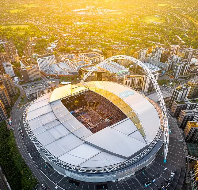 Aerial view of a large stadium with a distinctive arch and an open roof. The stands are filled with people, and a nearby cityscape is visible below a glowing sunset sky at a Taylor Swift concert.