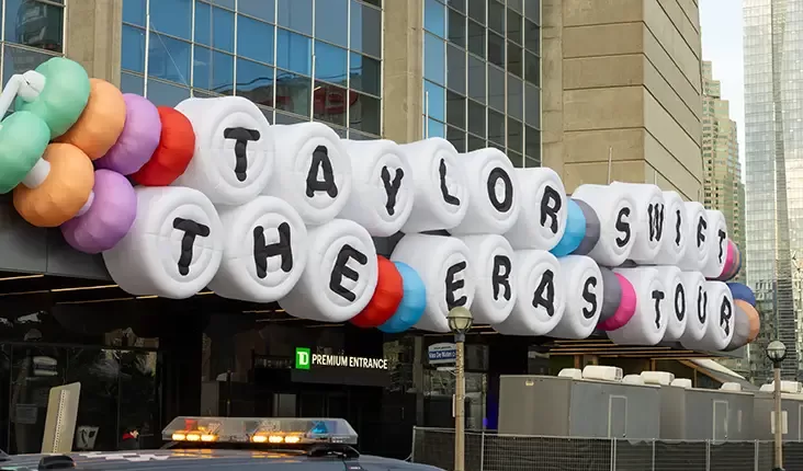 Large white balloons with black letters spell Taylor Swift The Eras Tour in front of a modern building. Colorful decorative balloons are attached on the sides, and a sign for a premium entrance is visible below.