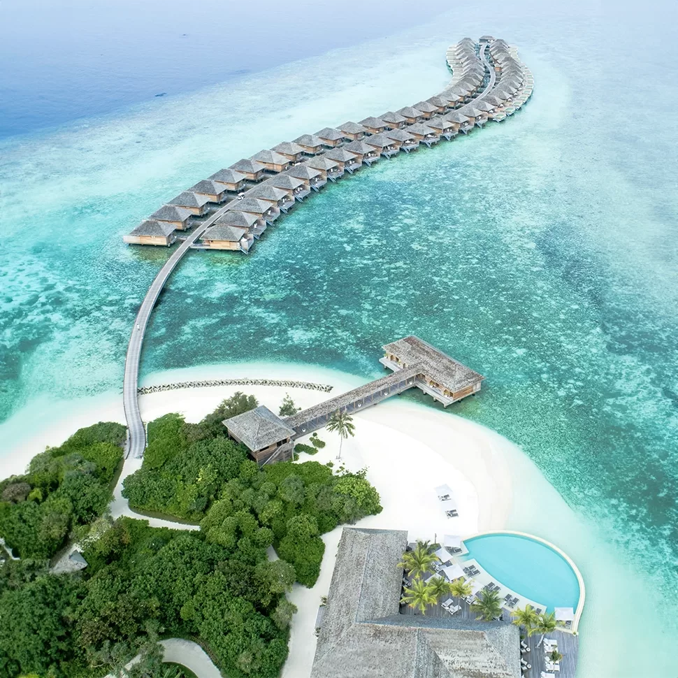 Aerial view of a luxury resort in the Maldives with overwater bungalows arranged in a curve above a turquoise lagoon, connected by wooden walkways.
