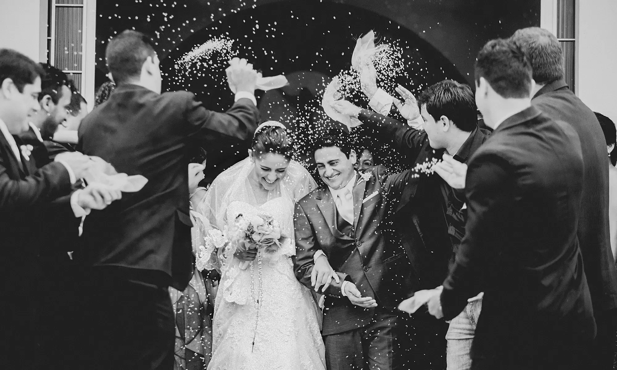 A bride and groom joyfully exit a building as guests shower them with rice and petals. The couple is smiling, holding hands, and dressed in wedding attire. Friends and family stand around them, celebrating. The scene captures a moment of happiness and festivity.