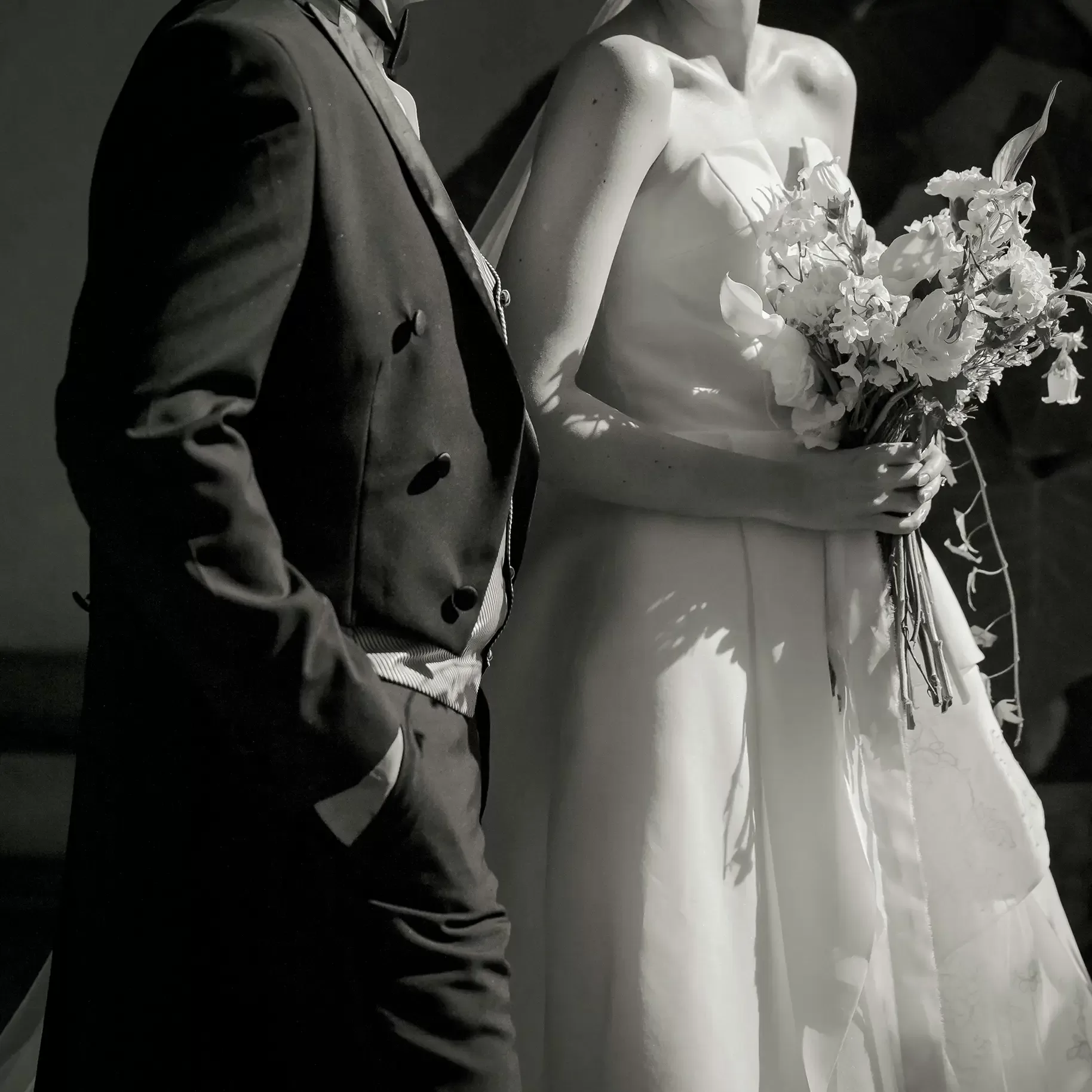 A black-and-white photo of a bride in a strapless wedding dress holding a bouquet of flowers, standing beside a person in a dark suit with one hand in their pocket. Their faces are not visible.