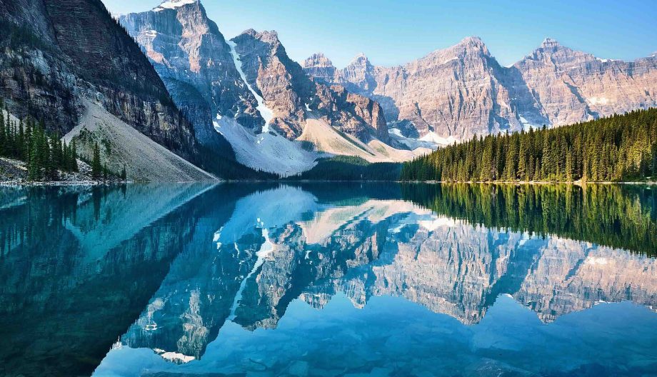 A tranquil lake reflects rugged, snow-capped mountains and dense coniferous forests under a clear blue sky. The shoreline is rocky, and the water is a striking blue-green, creating a stunning natural landscape.