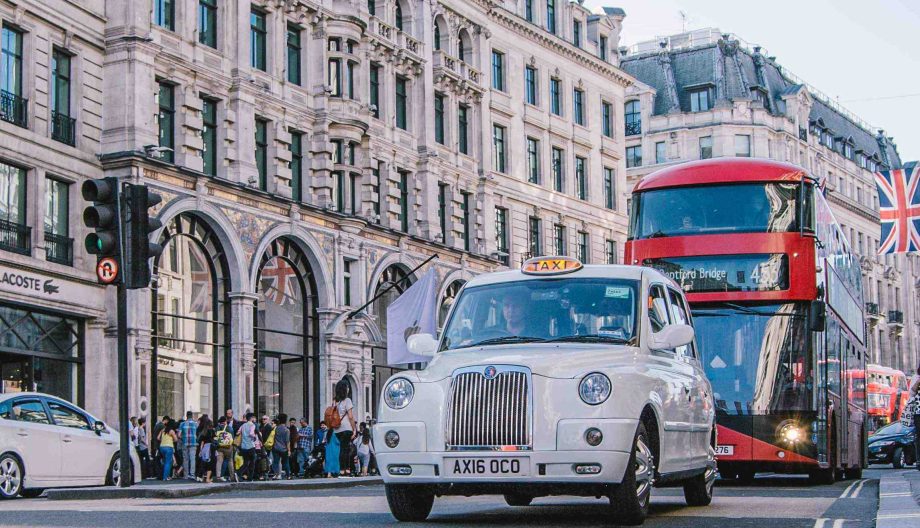 A white taxi and a red double-decker bus drive through a busy intersection in a city with historic architecture. Pedestrians walk on the sidewalk, and a British flag is visible in the background.