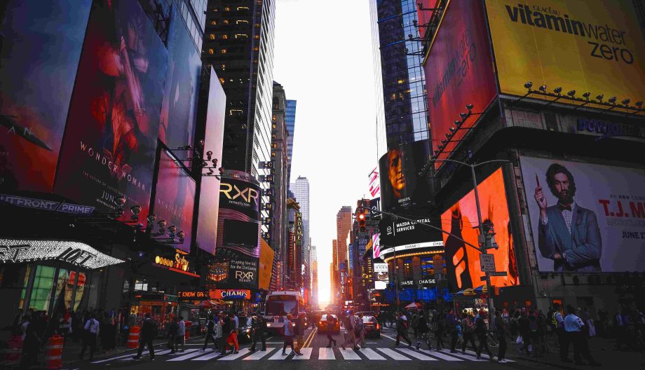 A vibrant city street scene at dusk with bright digital billboards and busy sidewalks filled with people. Cars are stopped at a crosswalk, and the sun sets between the tall buildings, casting a warm glow.