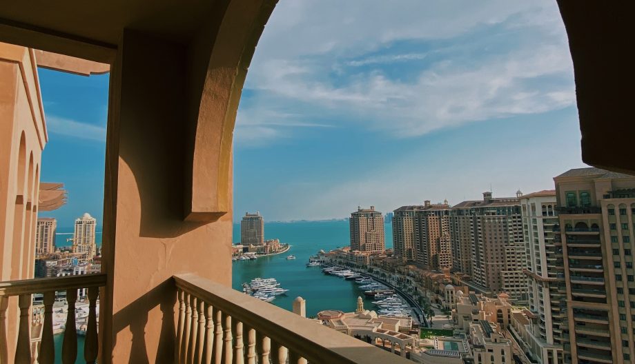 Scenic view from a balcony with an arched opening overlooking a coastal city. Tall buildings, a marina with boats, and a clear blue sky are visible in the background. The sunlight casts shadows on the balcony floor.