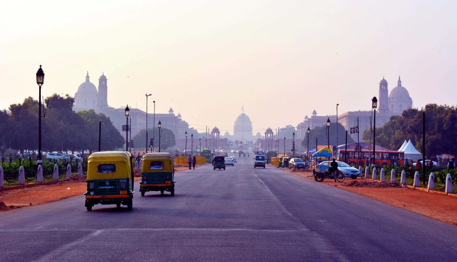 A bustling street in New Delhi with auto rickshaws and cars. The India Gate is visible in the background, framed by historical buildings and greenery on a hazy day.
