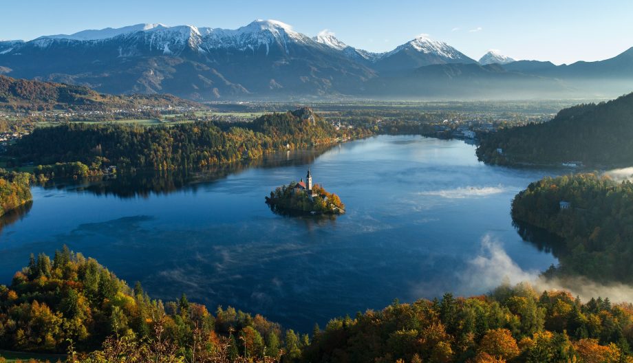 A serene lake surrounded by autumn-colored forests and mountains dusted with snow. In the lakes center, a small island features a church. Mist hovers above the water, and the morning light casts a warm glow over the landscape.