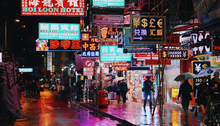 A vibrant city street at night with neon signs in various languages, including English and Chinese. Colorful advertisements for hotels and currency exchange are illuminated. People with umbrellas walk on the wet pavement.