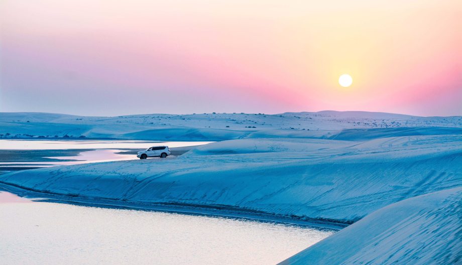 A white SUV parked on a vast, sandy desert landscape with shallow water pathways at sunset. The sky is a gradient of pink, orange, and blue hues, with the sun setting on the horizon.