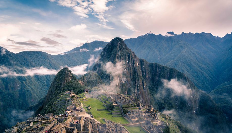A panoramic view of Machu Picchu in Peru, featuring ancient stone structures atop a lush green mountain. Mist lingers around the site, with towering peaks in the background under a partly cloudy sky.