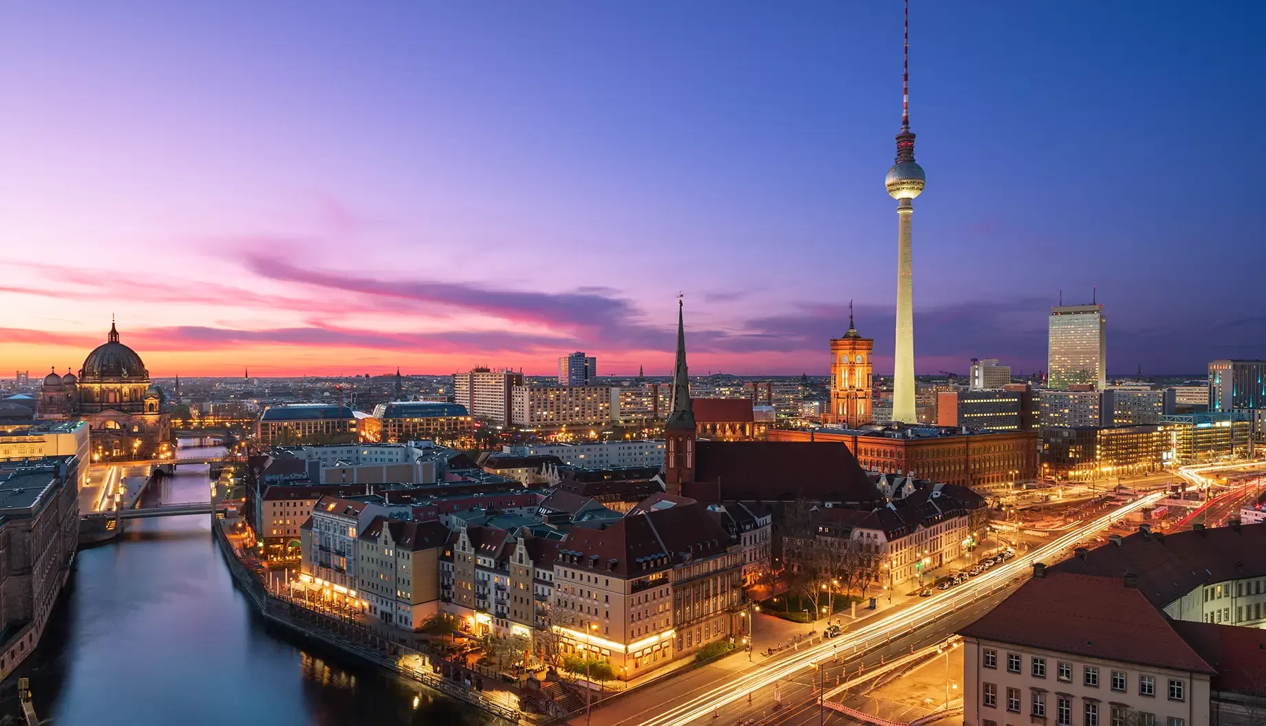A panoramic view of Berlin at twilight featuring the iconic Fernsehturm tower. The skyline is illuminated with city lights, buildings, and a river reflecting the vibrant colors of the sunset in the sky.