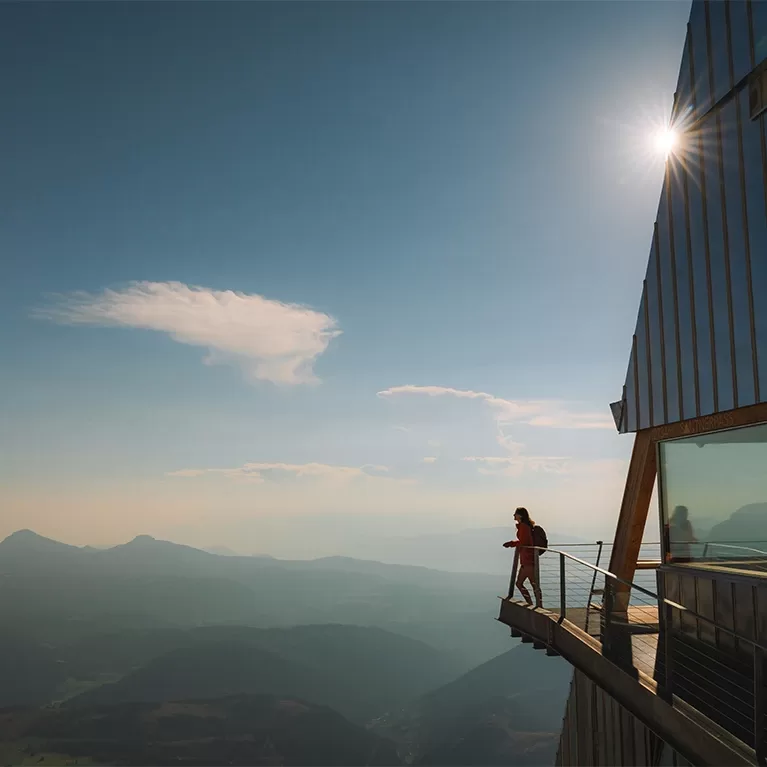 A man stands on a balcony, gazing at the majestic mountains in the distance, surrounded by a serene landscape.