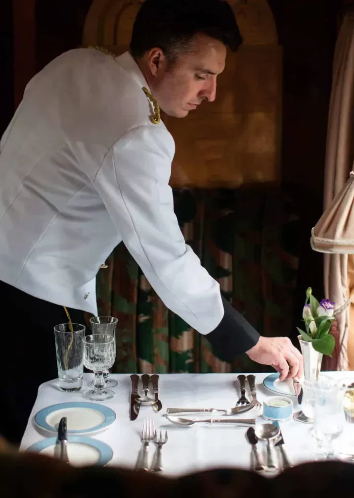 A man carefully arranging dishes and utensils on a dining table in preparation for a meal.