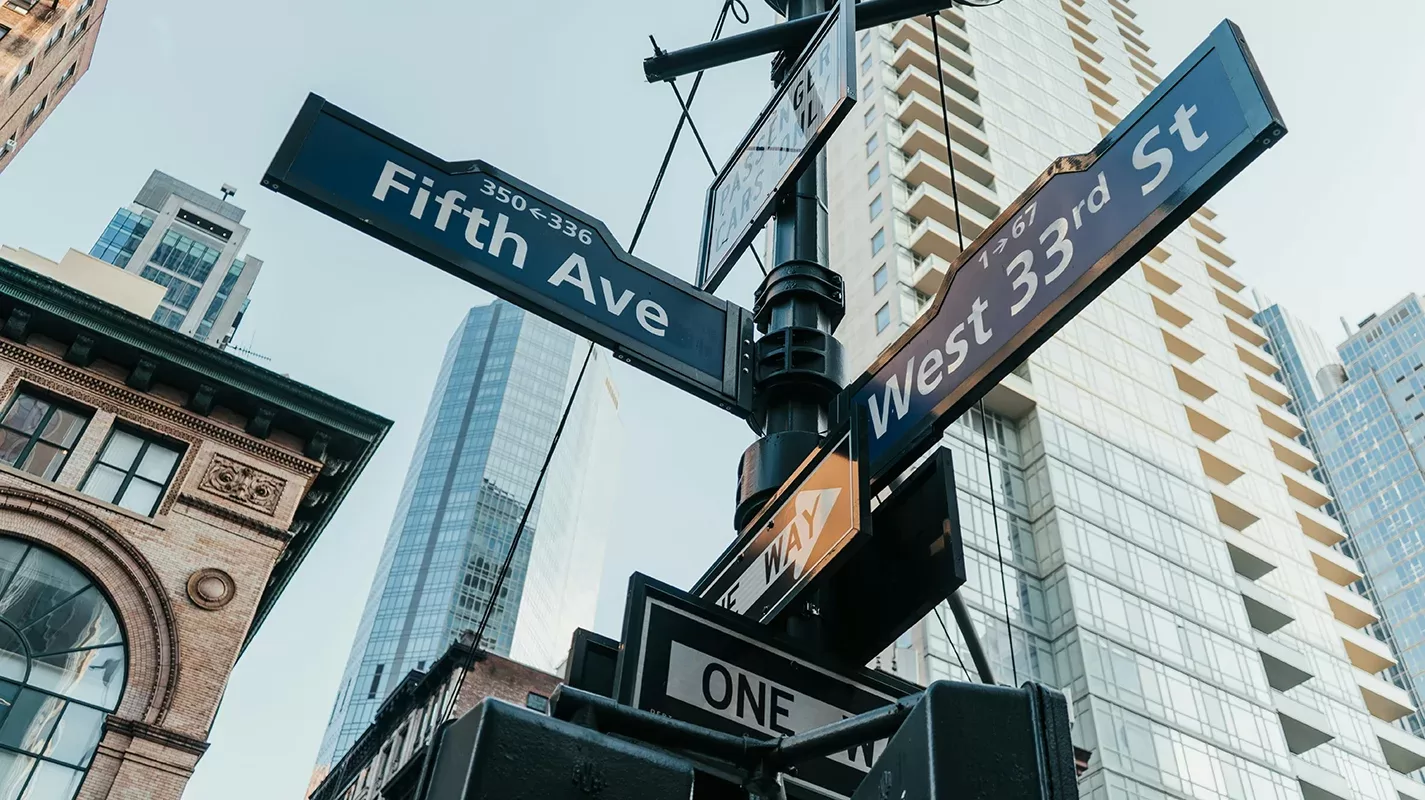Street signs at the intersection of Fifth Avenue and West 33rd Street in an urban area with tall buildings in the background, under a clear sky.