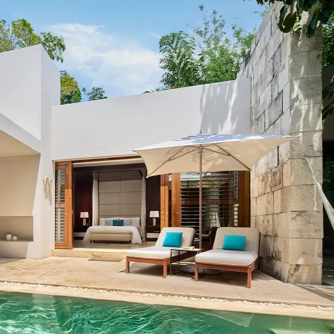 A modern villa with a private pool, two lounge chairs under a white umbrella, and an open sliding door revealing a bedroom with a neatly made bed and turquoise accent pillows. Lush greenery surrounds the area.