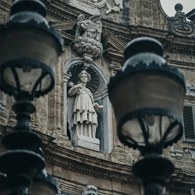 A stone statue of a robed figure stands in an ornate architectural niche on an old building, partially obscured by two large, vintage street lamps in the foreground.