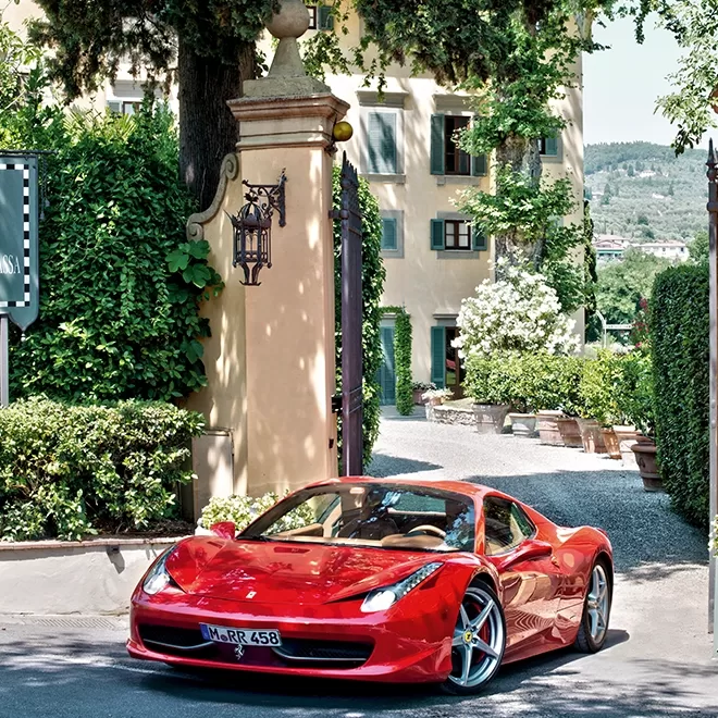 A red Ferrari sports car is parked on a driveway surrounded by greenery, with a large, elegant building and potted plants in the background under a sunny sky.