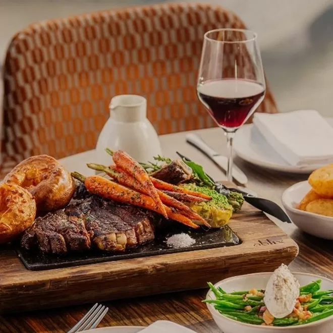 A wooden board with grilled steak, roasted carrots, broccoli, and Yorkshire puddings is set on a table next to a glass of red wine, a bowl of green beans, and a plate of more Yorkshire puddings.