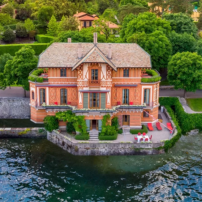 Aerial view of a large, elegant house with ornate woodwork and balconies, surrounded by lush greenery, situated directly on the edge of a lake with outdoor seating areas and gardens.