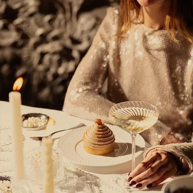 A woman in a sparkly beige top sits at a table with a dessert topped with swirled cream, a glass of white wine, and lit candles. The scene is warmly lit, creating an elegant and festive atmosphere.