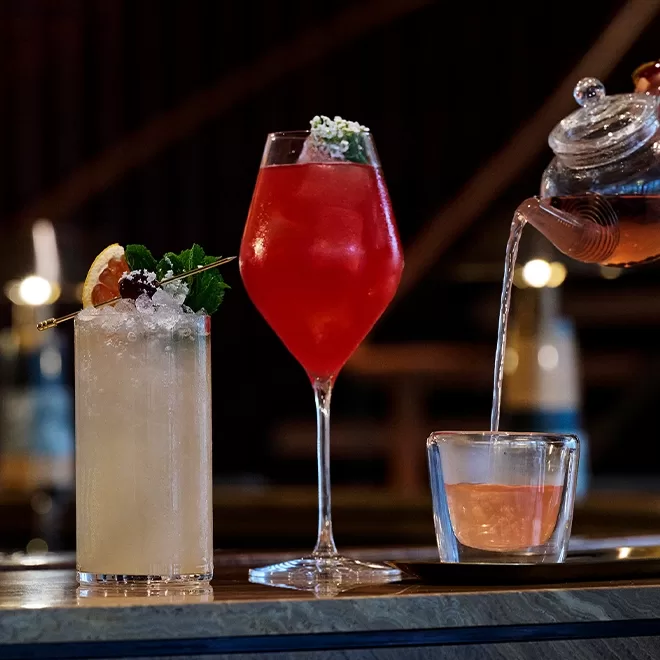 A yellow cocktail with crushed ice and garnish, a tall red drink in a wine glass with ice and flowers, and tea being poured from a teapot into a glass cup on a bar counter.
