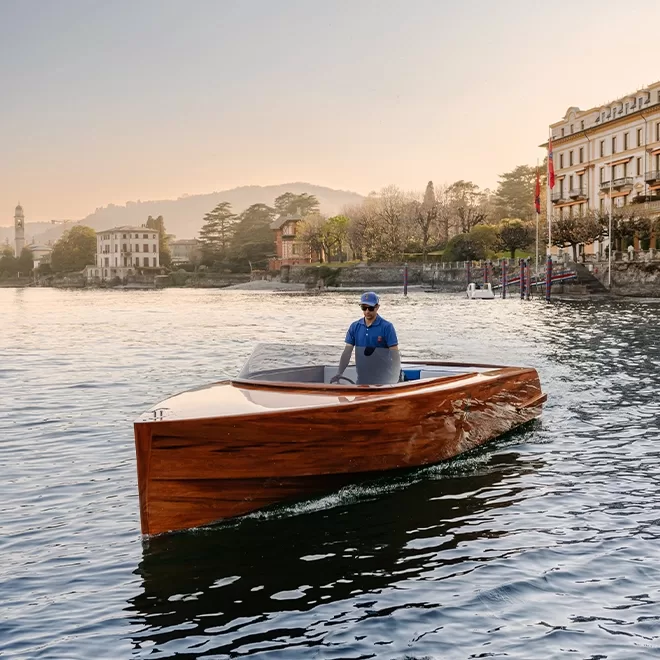A person steers a sleek, modern wooden boat on calm water near a shoreline lined with elegant buildings and trees under a soft evening sky.