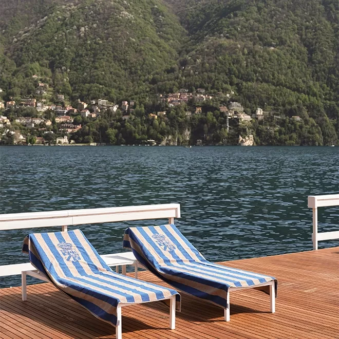 Two striped blue and white lounge chairs sit on a wooden deck overlooking a calm lake, with green, forested hills and houses visible in the background.