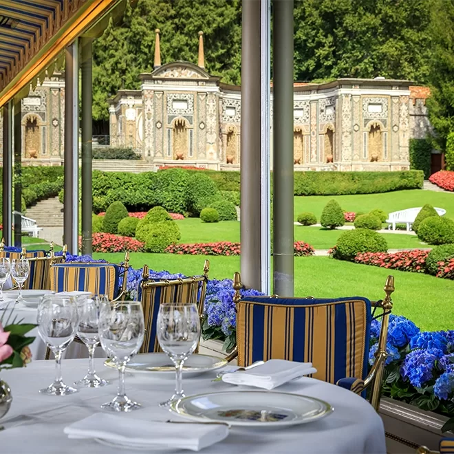 Elegant outdoor dining area with striped chairs and set tables overlooks a manicured garden with colorful flowers, trimmed bushes, and a grand stone building in the background.