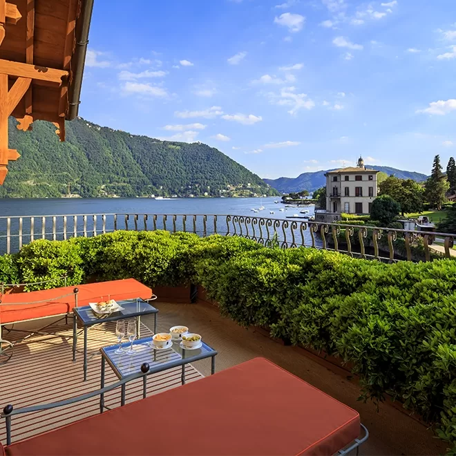 A scenic terrace with orange lounge chairs and small tables overlooks a lake surrounded by green mountains, with a historic building and lush gardens in the background under a blue sky.