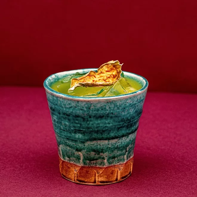 A green cocktail with ice and a dried citrus garnish is served in a textured blue and brown ceramic cup, set against a deep red background.