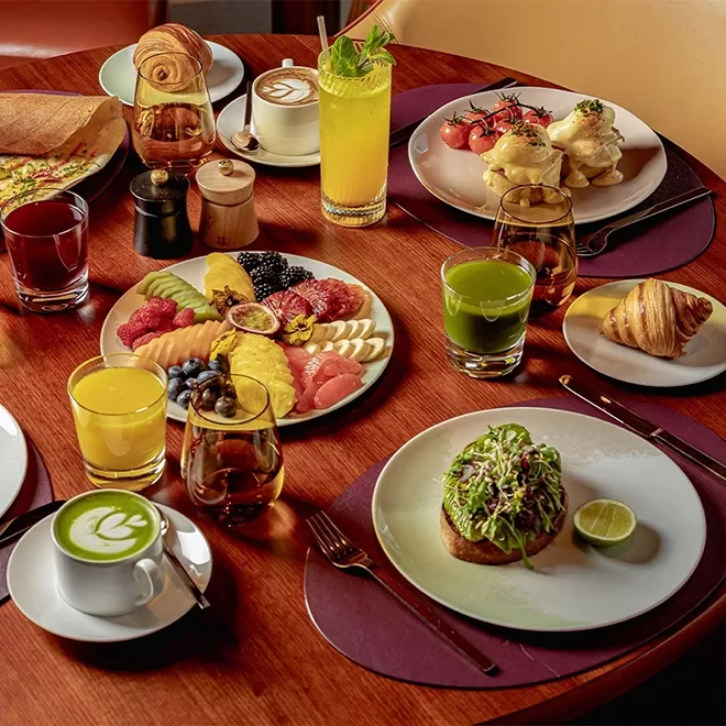 A wooden table set for breakfast with plates of fresh fruit, avocado toast, eggs benedict, pastries, green juice, orange juice, coffee with latte art, and a glass of water.