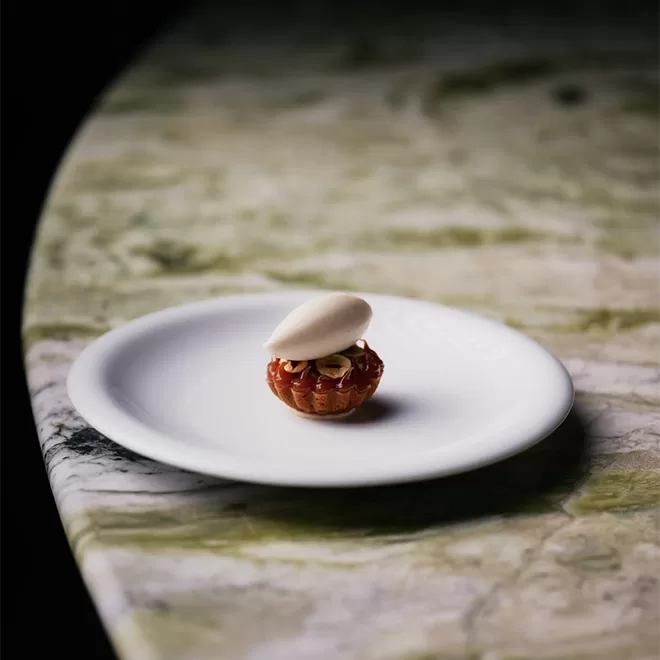 A small tart topped with a scoop of ice cream sits on a white plate, placed on a marbled green and beige table with a softly focused background.